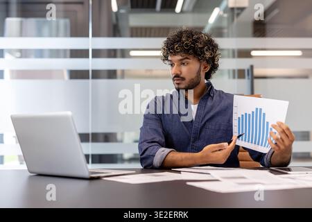 Young Indian man sitting at a desk in the office, talking on a video call and showing graphs on the laptop camera. Stock Photo
