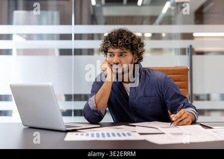 Young Indian businessman sitting at his desk in the office, looking bored and tired at his laptop. Stock Photo