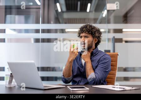 A young Indian man sits at a desk in an office, holding his throat with his hand and using pain reliever. Stock Photo