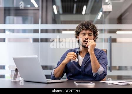 Sick young Indian man sitting at a desk in the office, holding a tissue in his hand and using a nasal spray. Stock Photo