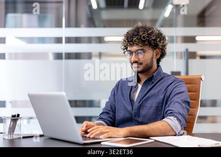 Concentrated young Indian man wearing glasses sitting at a desk in the office and working on a laptop. Stock Photo