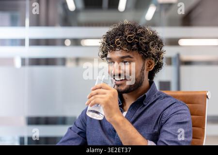 Close-up photo of a young Indian man sitting in an office and drinking water from a glass. Stock Photo