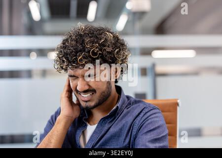 Close-up photo of an Indian man indoors, holding his ear with his hand, grimacing in pain. Stock Photo