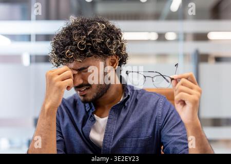 Close-up photo of a tired young Indian man in the office, holding his glasses in his hand and rubbing his eyes. Stock Photo