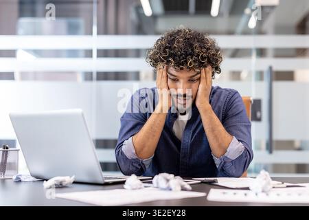 Upset and tired Indian man sitting at office desk, holding head in hands and looking at crumpled paper on table. Stock Photo
