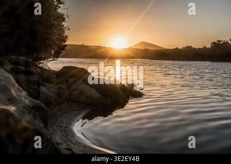 Evening sun over a tranquil lake with rocky shore and silhouetted trees. Stock Photo