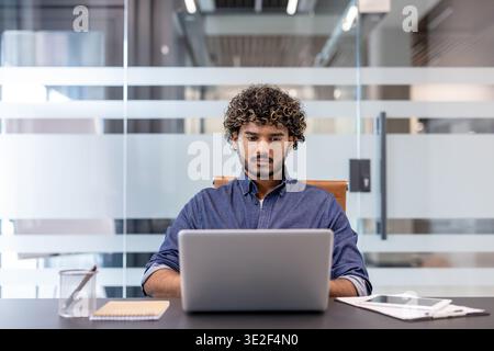 Serious and confused young Indian man sitting at a desk in the office and working on a laptop. Stock Photo