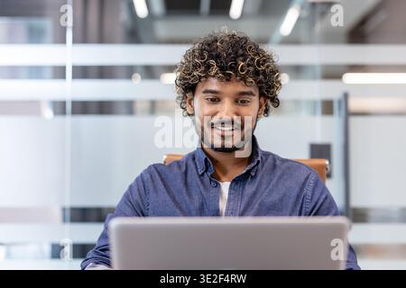 Close-up photo of a young Indian man smiling while working on a laptop in the office. Stock Photo