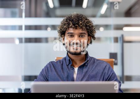 Close-up portrait of a smiling young Indian man sitting in the office at his workplace and laptop and looking at the camera. Stock Photo