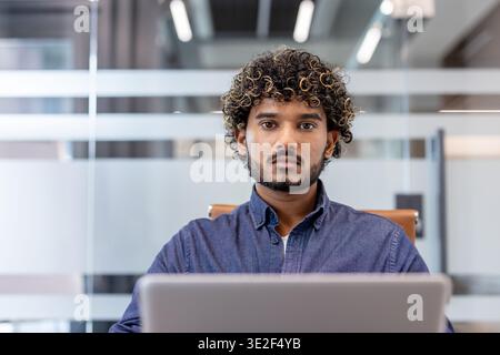 Close-up photo of a young Indian businessman sitting at a laptop in the office and looking seriously at the camera. Stock Photo