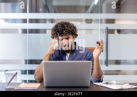 Upset young Indian man sitting at a desk in the office, looking at the laptop screen in frustration and holding his head. Stock Photo