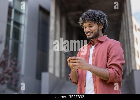 Smiling young Indian man in a checkered shirt stands on the street and uses a mobile phone he holds in his hands. Stock Photo