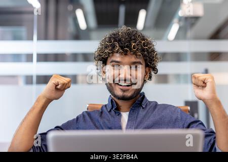 Close-up photo of a young happy smiling Indian man sitting at a desk in the office with a laptop and showing a sign of strength and success with his hands at the camera. Stock Photo