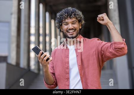 Close-up portrait of a young Indian man holding a phone in his hand and showing a victory gesture with his hand. Stock Photo