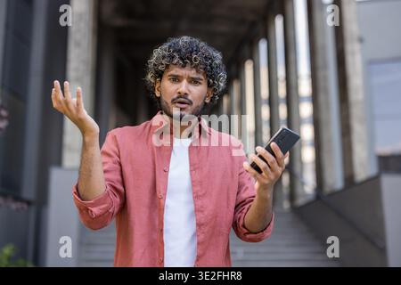 Worried young Indian man standing outside, holding phone, spreading hands in frustration and looking at camera. Stock Photo