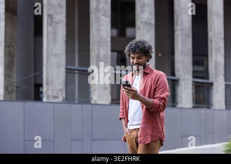 Smiling young Indian man walking down a city street, holding his hand in his pocket and using his mobile phone. Stock Photo