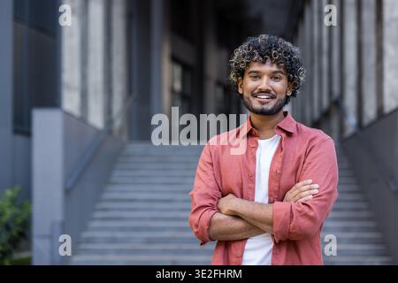 Portrait of a young Indian man in a red shirt standing smiling near an office building, arms crossed over his chest and smiling at the camera. Stock Photo