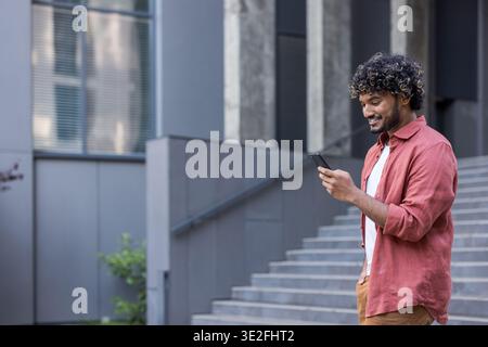 A young Muslim man stands to the right of an office building in a white shirt and uses his mobile phone with a smile. Stock Photo