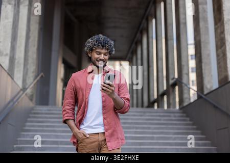 A young Indian man is walking down the stairs of an office building, holding his left hand in his pocket and looking at his mobile phone screen. Stock Photo