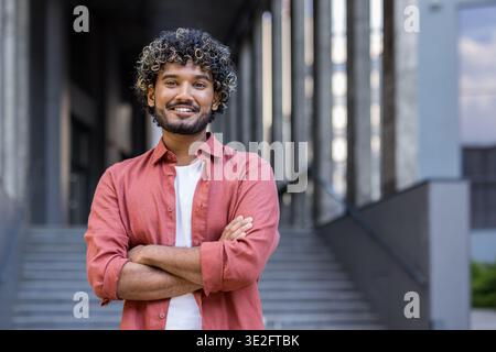 Portrait of a young Indian man in a red shirt standing on the street, arms crossed on his chest and looking at the camera with a smile. Stock Photo