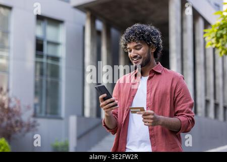 Young Indian man standing on the street, using mobile phone and credit card. Stock Photo