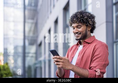 Close-up photo of a smiling young Indian man standing outside and using his mobile phone. Stock Photo