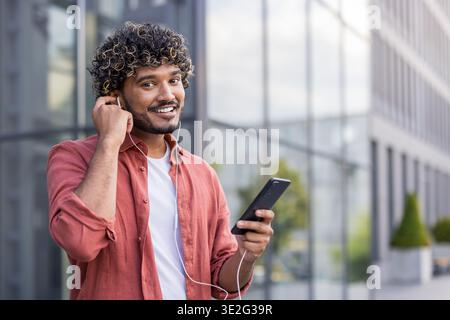 Close-up portrait of a young Indian man sitting outside wearing headphones holding a phone and smiling at the camera. Stock Photo