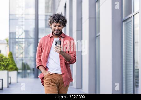 Smiling young Indian man walking down a city street, holding his hand in his pocket and using his mobile phone. Stock Photo