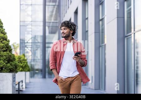 A young Indian man in a red shirt is walking down the street, holding his hand in his pocket, using his phone, and looking to the side with a smile. Stock Photo
