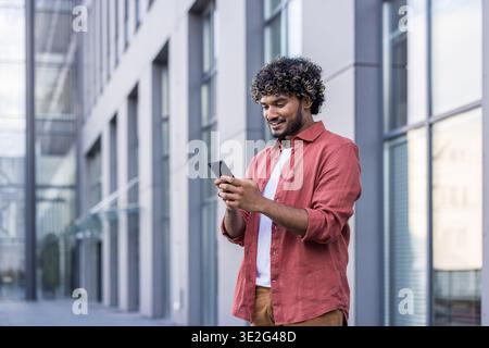 Smiling young Indian man standing outside a building and using his mobile phone. Stock Photo