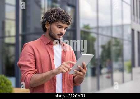 Close-up photo of a young Indian man standing outside and using a tablet. Stock Photo