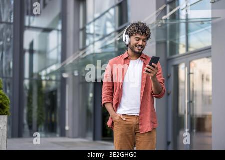 An Indian young man wearing headphones walks through the city and uses a mobile phone. Stock Photo