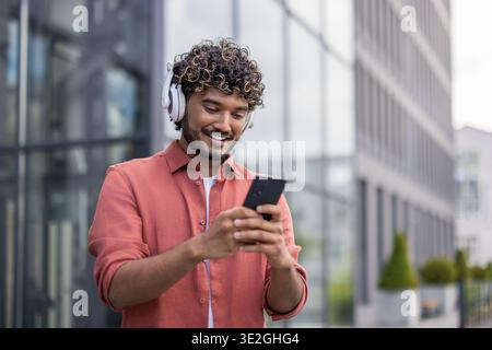 Close-up photo of a young Indian man wearing headphones standing outside and using his phone. Stock Photo