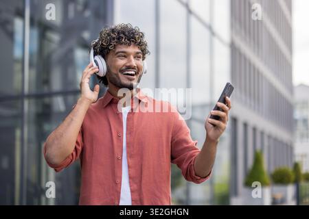 Smiling and happy young Indian man wearing headphones, holding phone, dancing and listening to music. Stock Photo