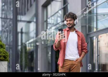 Young Indian man wearing headphones walking on the street and using his mobile phone. Stock Photo