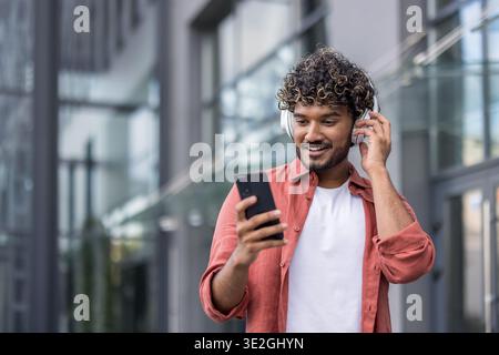 Indian smiling young man wearing headphones and shirt is outside using mobile phone. Stock Photo
