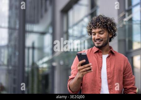 Close-up photo of a young Indian man standing on the street with a smile and using a mobile phone. Stock Photo