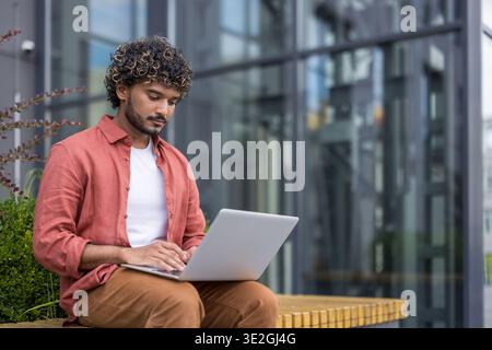 Serious young Indian man sitting on a bench outside, working and studying on a laptop. Stock Photo