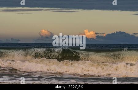Waves crash on a sandy beach under a warm sunset sky, creating a dramatic coastal scene. Stock Photo