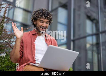 Smiling young Indian man wearing headphones sitting on a bench outside with a laptop on his lap and talking on a video call. Stock Photo