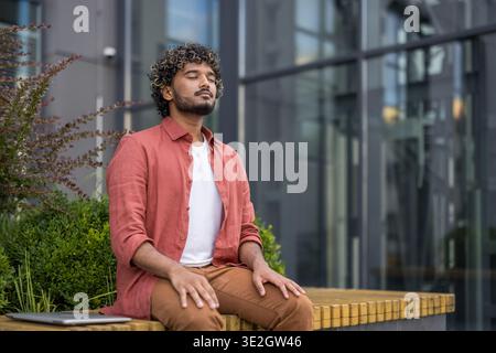 A young Indian man sits on a bench outside near a building with his eyes closed and resting relaxed. Stock Photo