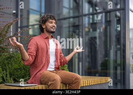 Smiling and relaxed young Indian man sitting on a bench near a building in a yoga lotus pose and meditating. Stock Photo