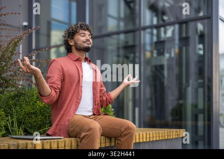 Relaxed young Indian man sitting on a bench outside a building in lotus position with eyes closed and meditating. Stock Photo