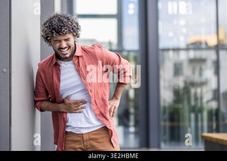 A young Indian man stands outside, leaning against a wall, holding his stomach in his hand, suffering from severe pain. Stock Photo