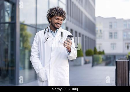 Smiling young Indian male doctor standing in white coat on the street and using mobile phone. Stock Photo