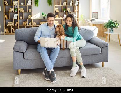 Happy couple with dog using laptop at home on sofa, remote work and leisure Stock Photo