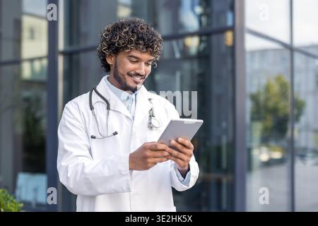 Close-up photo of a young Indian male doctor standing outside a hospital and using a tablet in his hands. Stock Photo