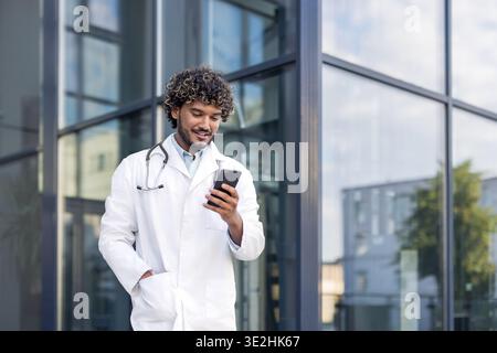 Smiling Indian man doctor standing in white uniform on the street and using mobile phone. Stock Photo