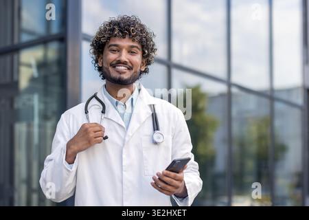 Close-up portrait of a young Indian doctor in a white coat standing outside, holding a phone and a stethoscope, smiling and looking at the camera. Stock Photo