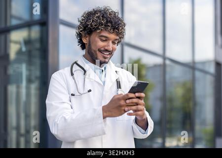 Close-up photo of a young Indian male doctor in a white coat standing near a hospital and using a mobile phone. Stock Photo